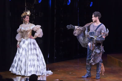 Kate in silvery ball gown and crown-like hat with left hand on hip, Petruchio in oeralls, t-shirt and holding his antler helmet and tarp for a cloak as he reaches his gloved hand to her