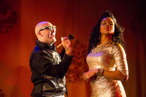 The makeup artist is bald, wearing black leather jacket, and black glasses holding up a brush to Rosalind in a glittering gold, tight dress, jeweled bracelet on her rist, and hair cascading down, her head turned and eyes closed.