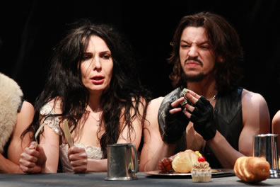 Kate, knife and fork in hand, is denied food by Petruchio, who is eating at a banquet table