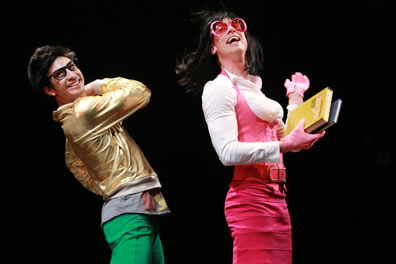Tranio in gold jacket and green pants with Lucentio desguised as a woman tutor in a tight-fitting white sweater and red halter dress and oversized pink sunglasses