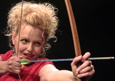 Beckman wearing red dress aims a bow and arrow to the right