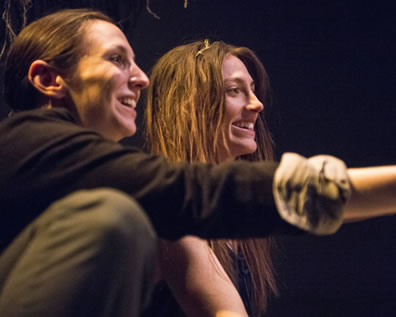 Ferdinand in black sweater, rolled up sleeves, and green pants smiling and pointing with Miranda, also smiling, in a simple dress hard to see with her long stringy hair ad bare arm; both are looking off to the right of the picture.