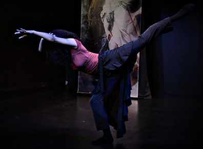 Martinez de Osaba in blue paints, blue dress, and red top pauses bent over at the waist, standing on her right foot, her left foot extended high behind her and her left arm, grasped by her right hand, extended out in fron of her lowered head. 