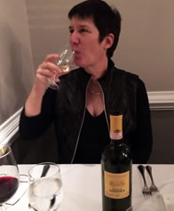 Sarah in a sexy black dress with high colar jacket sipping wine at a restaurant table